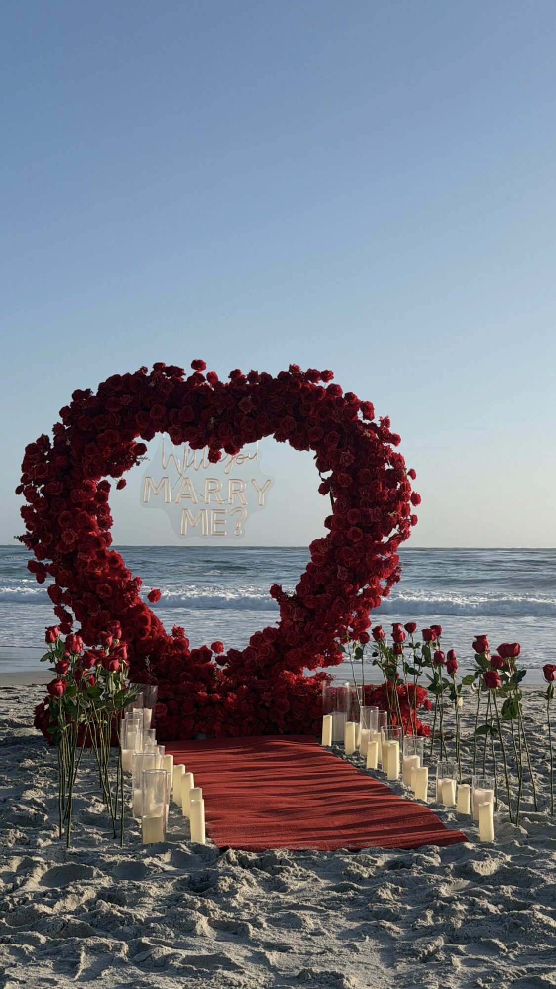 Heart-shaped floral arch on beach with candles and red carpet, ocean and sky in background.