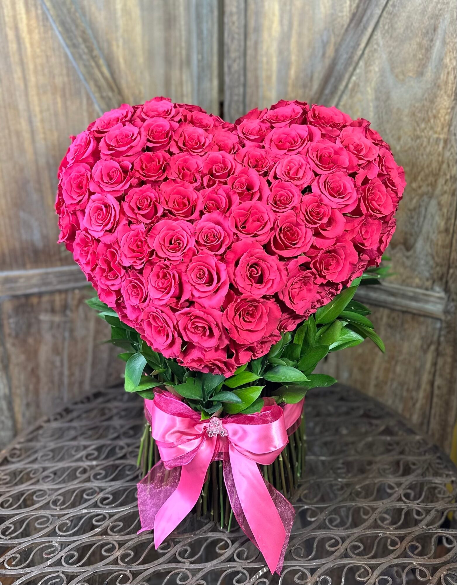 Heart-shaped arrangement of pink roses in a glass vase with green leaves and pink ribbon, on a wooden surface.