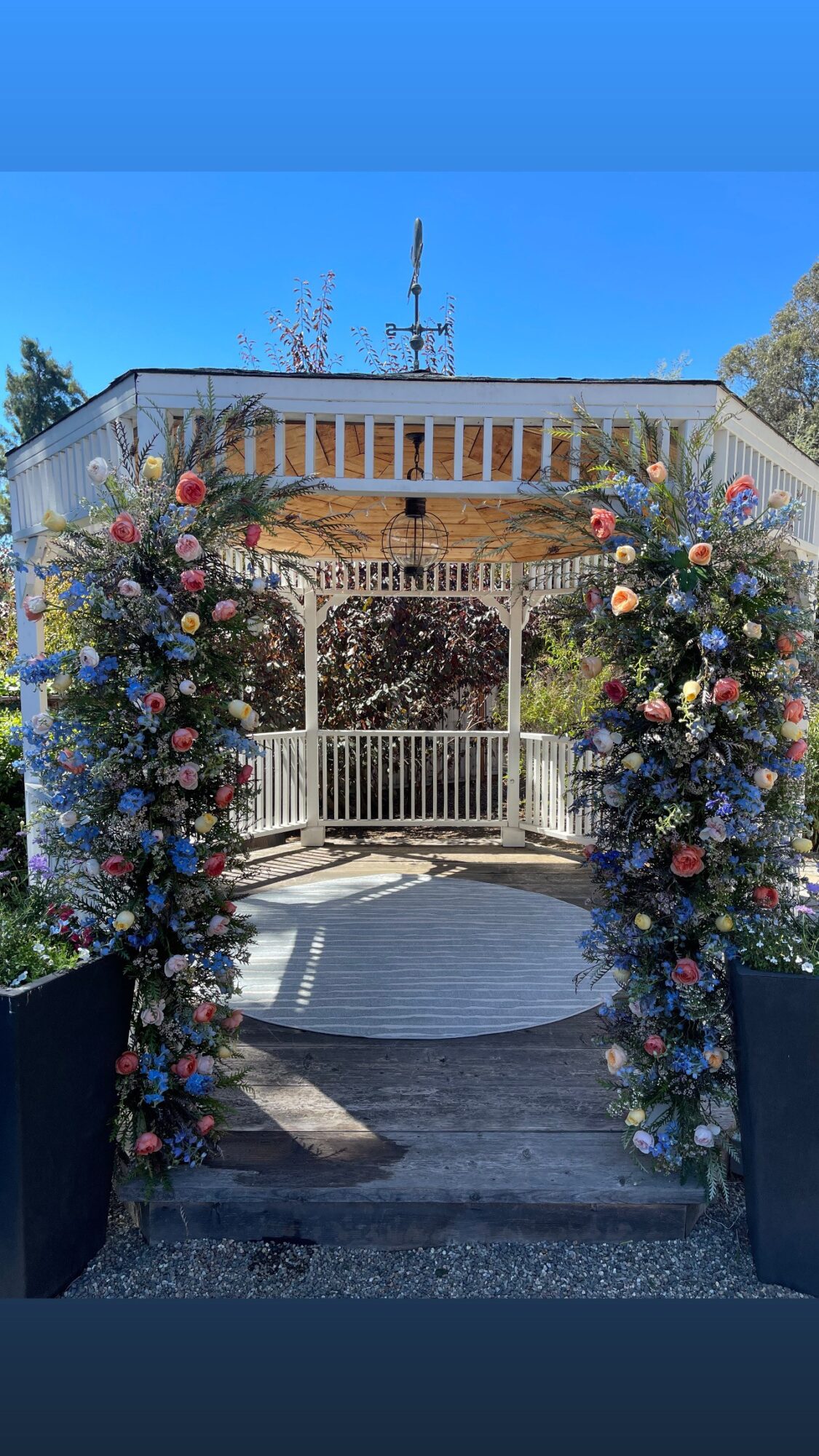 Decorated archway with colorful flowers and greenery, leading to a white fence and a building with a balcony, under a blue sky.