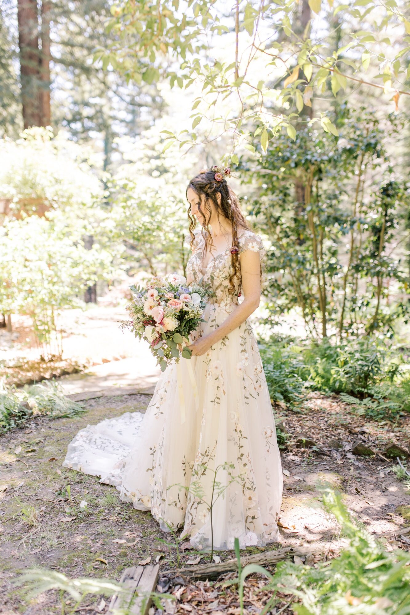 Woman in a wedding dress holding a bouquet in a sunlit garden surrounded by trees and greenery.