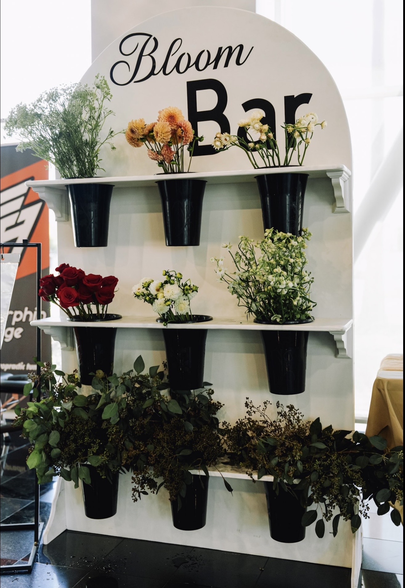 Display shelf with potted flowers and a sign reading 'Bloom Bar' in a bright room.