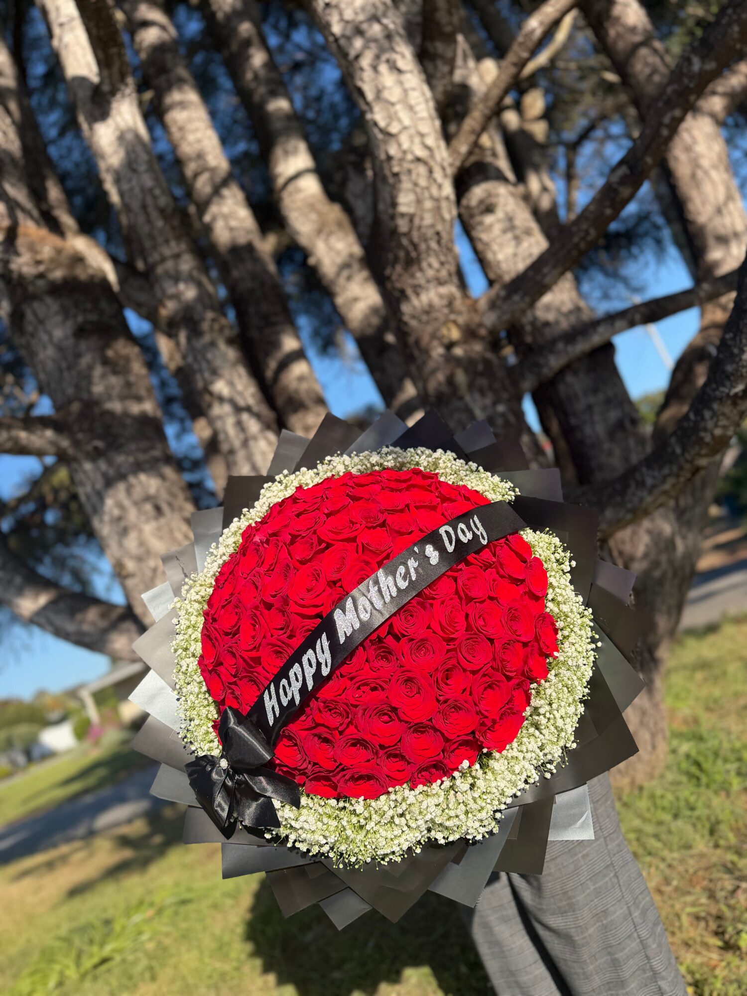 Bouquet of red roses with white baby's breath, wrapped in black paper, with a black ribbon reading 'Happy Mother’s Day'.