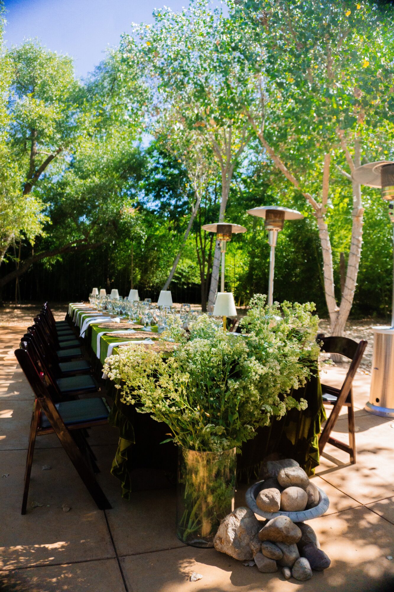 Outdoor dining table with chairs, surrounded by trees and greenery, with a centerpiece of flowers and stones, under sunlight.