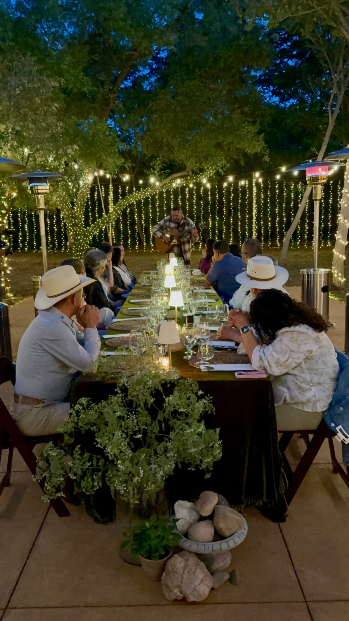 People sitting at a long outdoor table with string lights and trees in the background, some wearing hats, during evening.