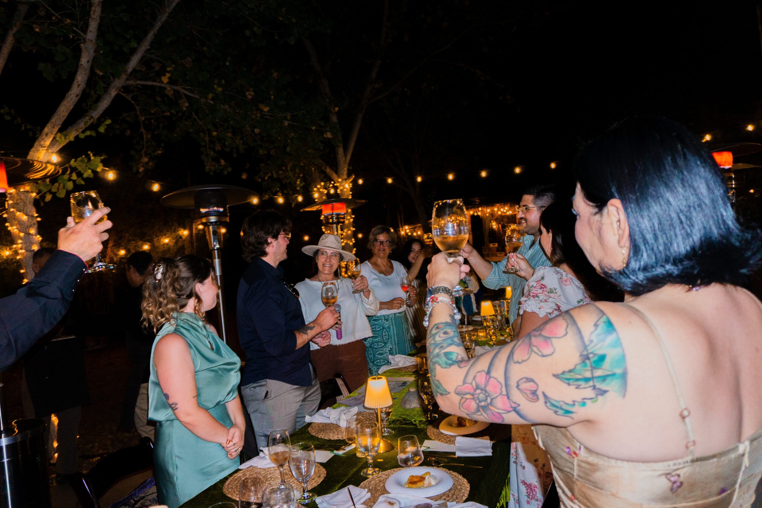 Group of people raising glasses in celebration at an outdoor nighttime event with string lights and decorated tables.