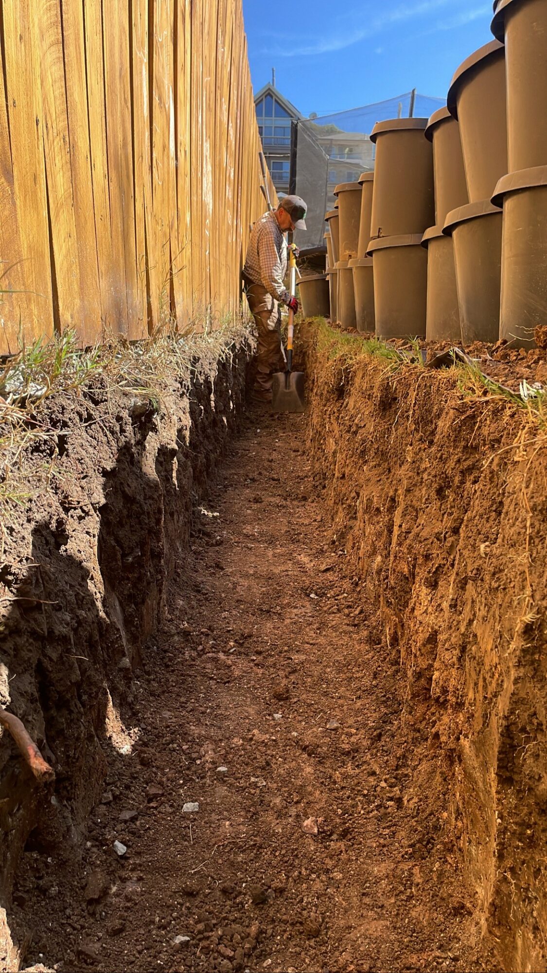 A person working in a deep trench between a wooden fence and large concrete pipes, with a cityscape in the background.