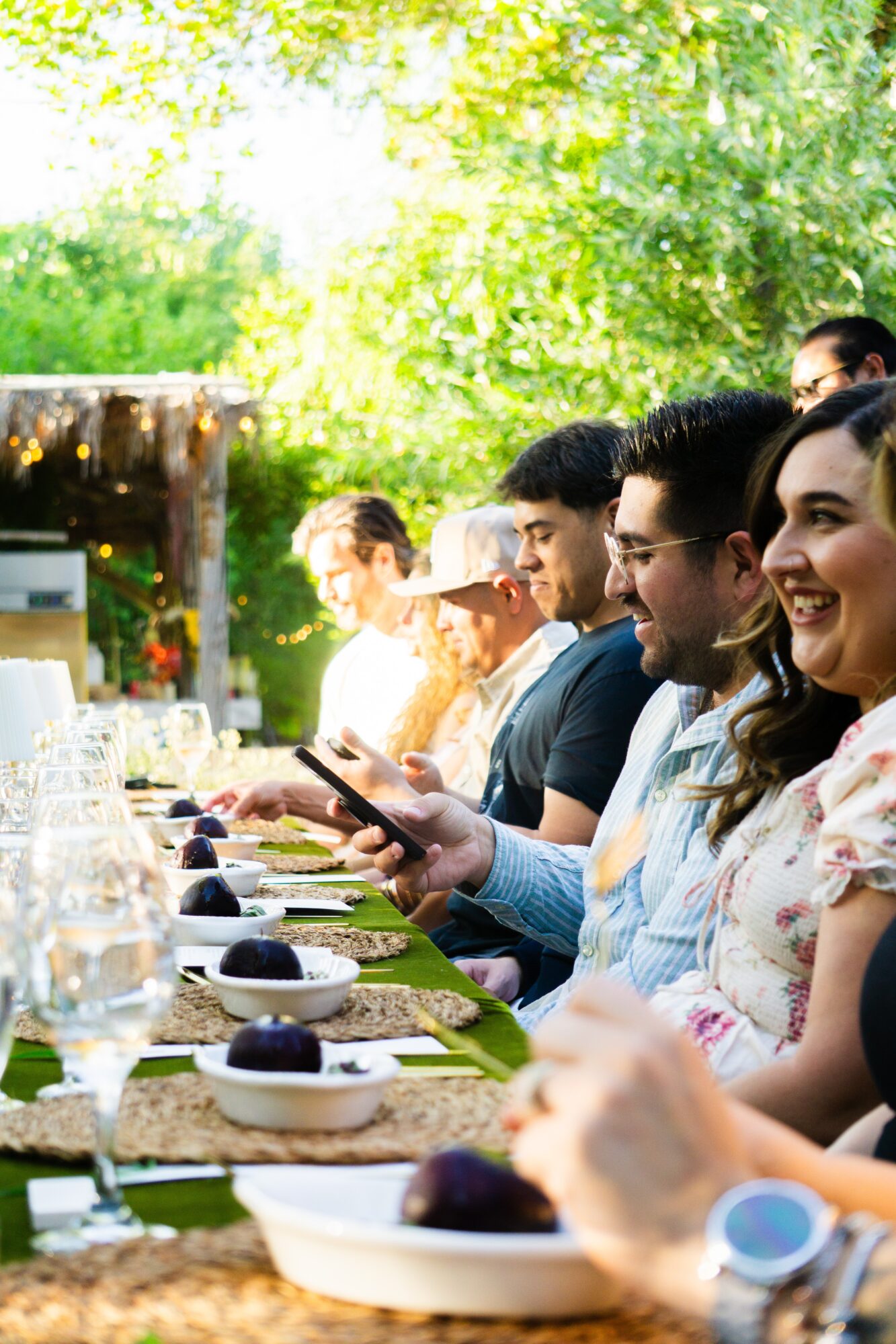 People sitting at a long outdoor table with food and drinks, smiling and engaging, surrounded by green trees.