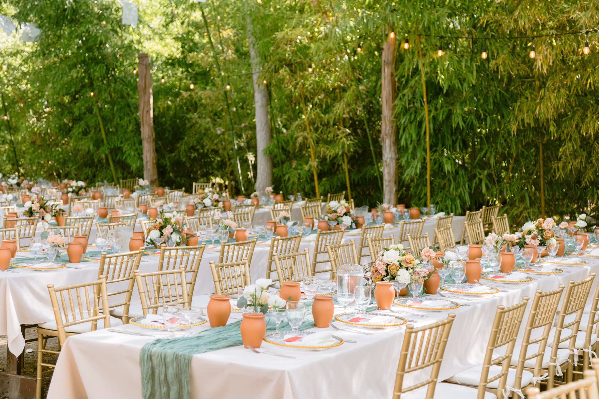Outdoor event with long tables, white tablecloths, floral centerpieces, and chairs, set against a green wooded background.