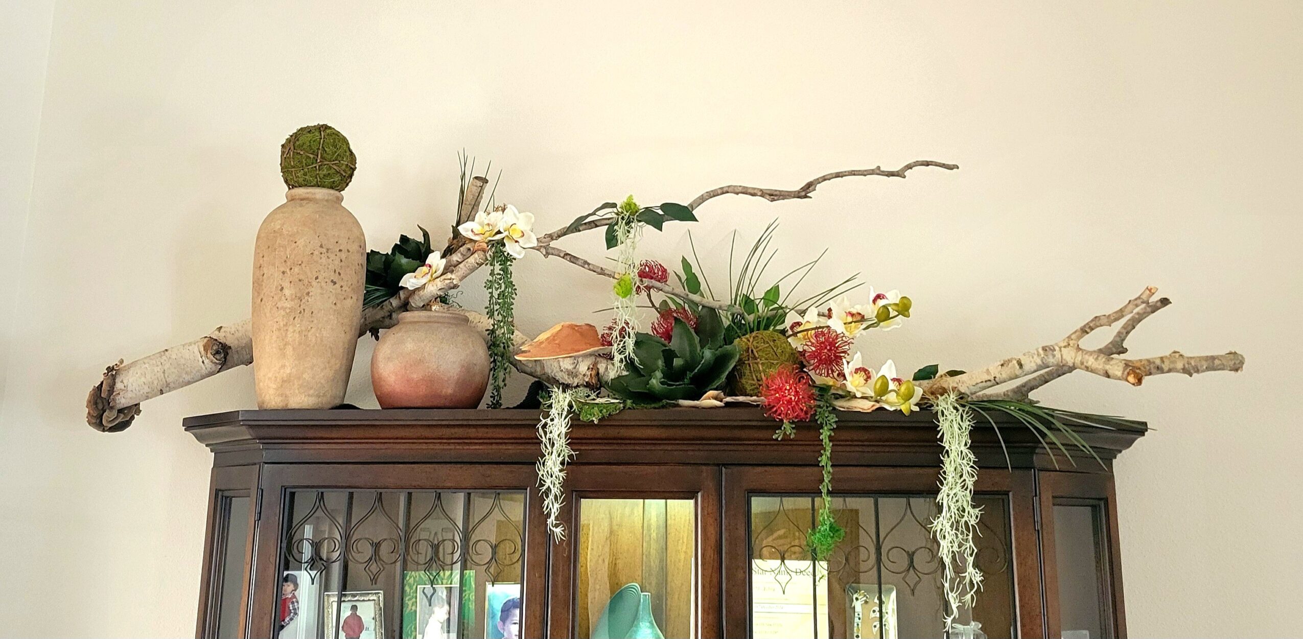 Decorative arrangement with vases, flowers, and branches on top of a wooden cabinet, against a plain wall.