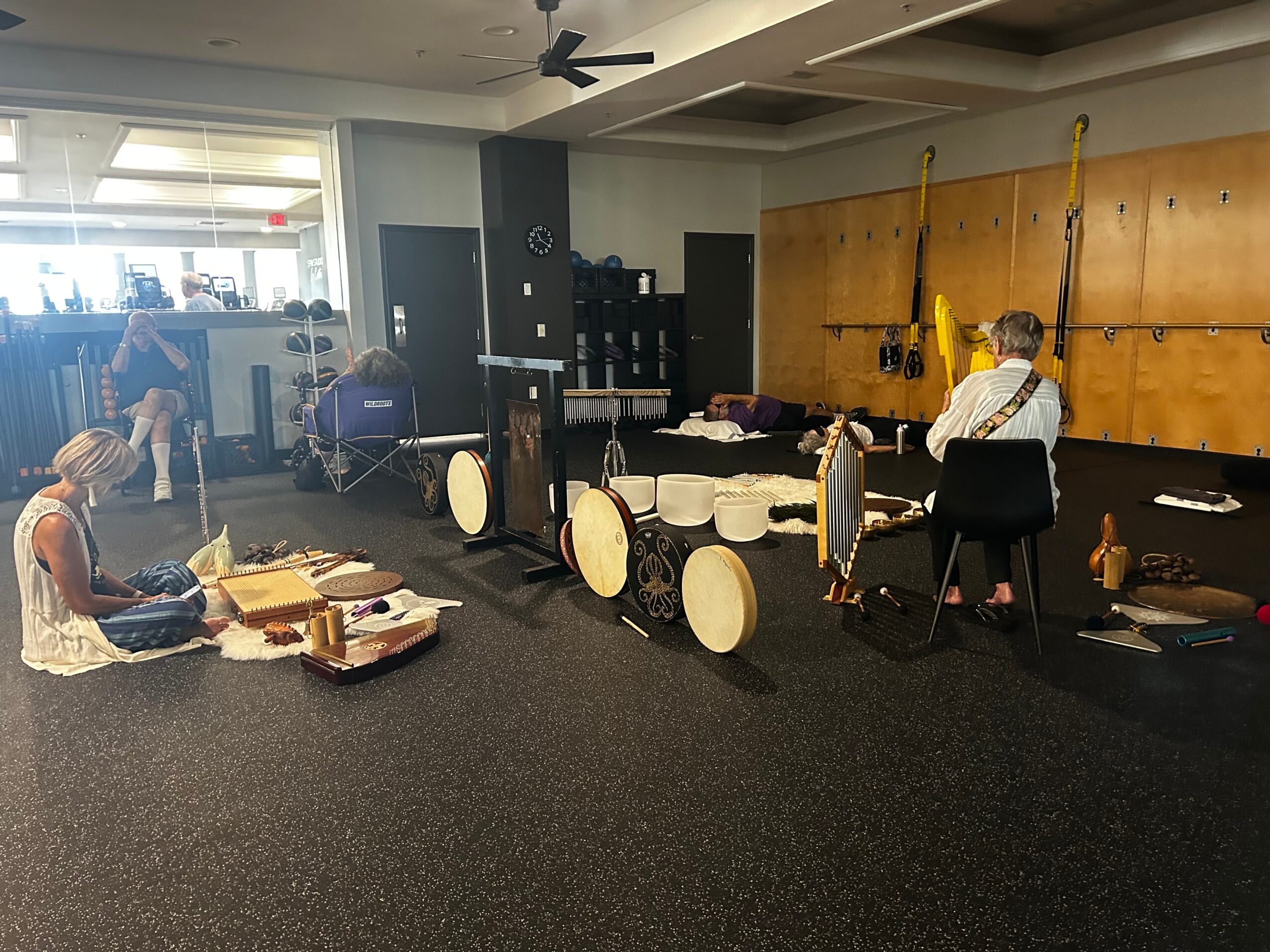 Person sitting on the floor with boxes, musical instruments and equipment nearby, in a room with a wooden wall and exercise equipment.