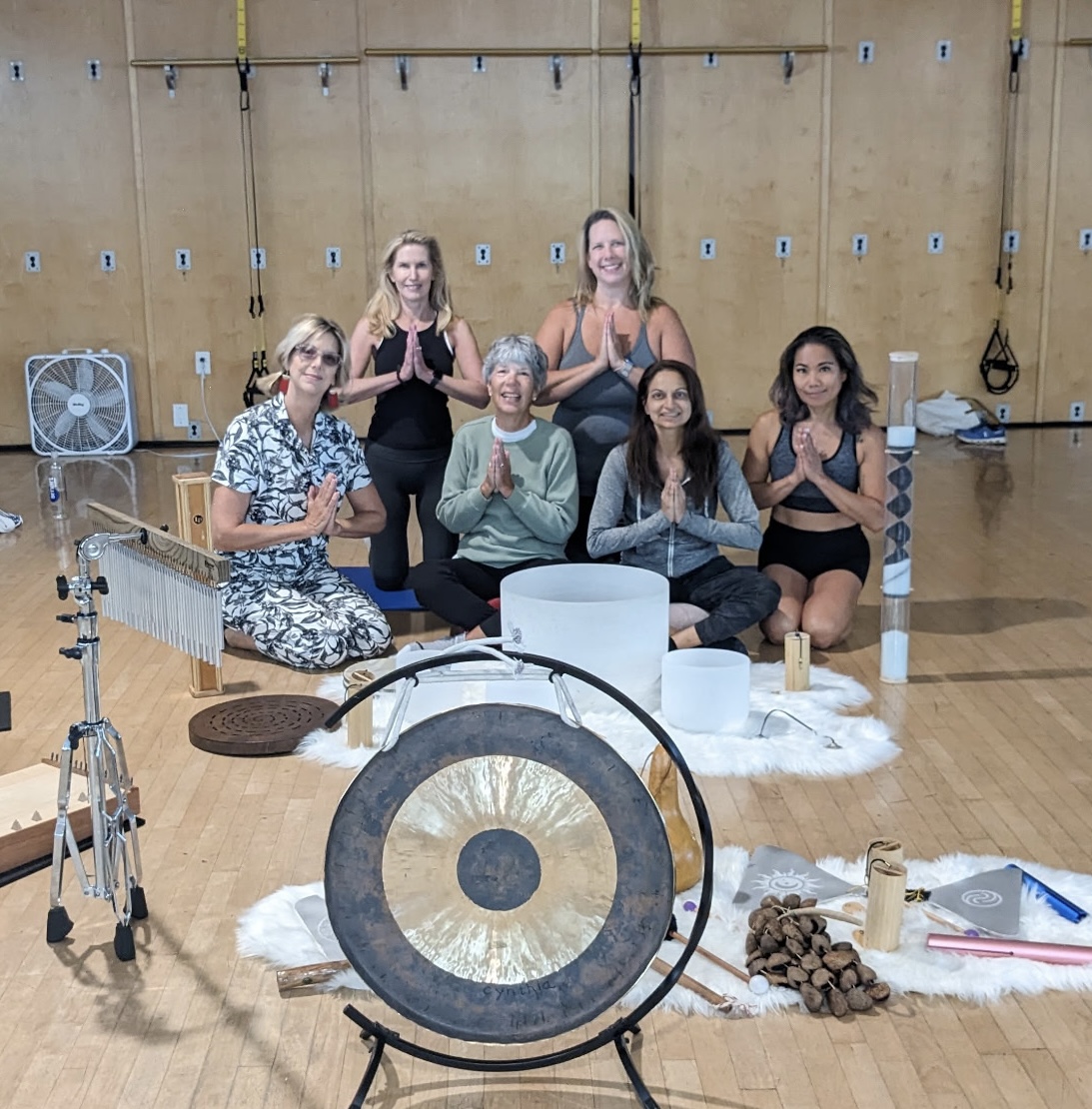 Six women sitting and standing in a yoga studio, meditating with hands in prayer position, surrounded by yoga props and equipment.