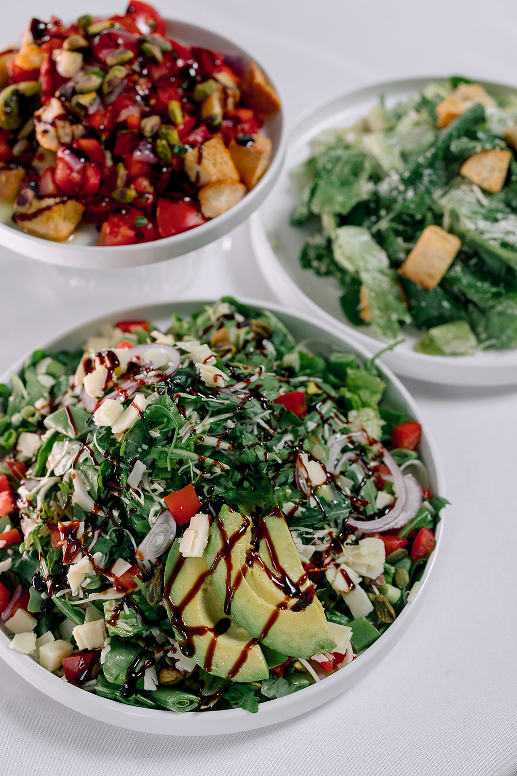 Three bowls of colorful salads with vegetables, greens, and toppings on a white surface.