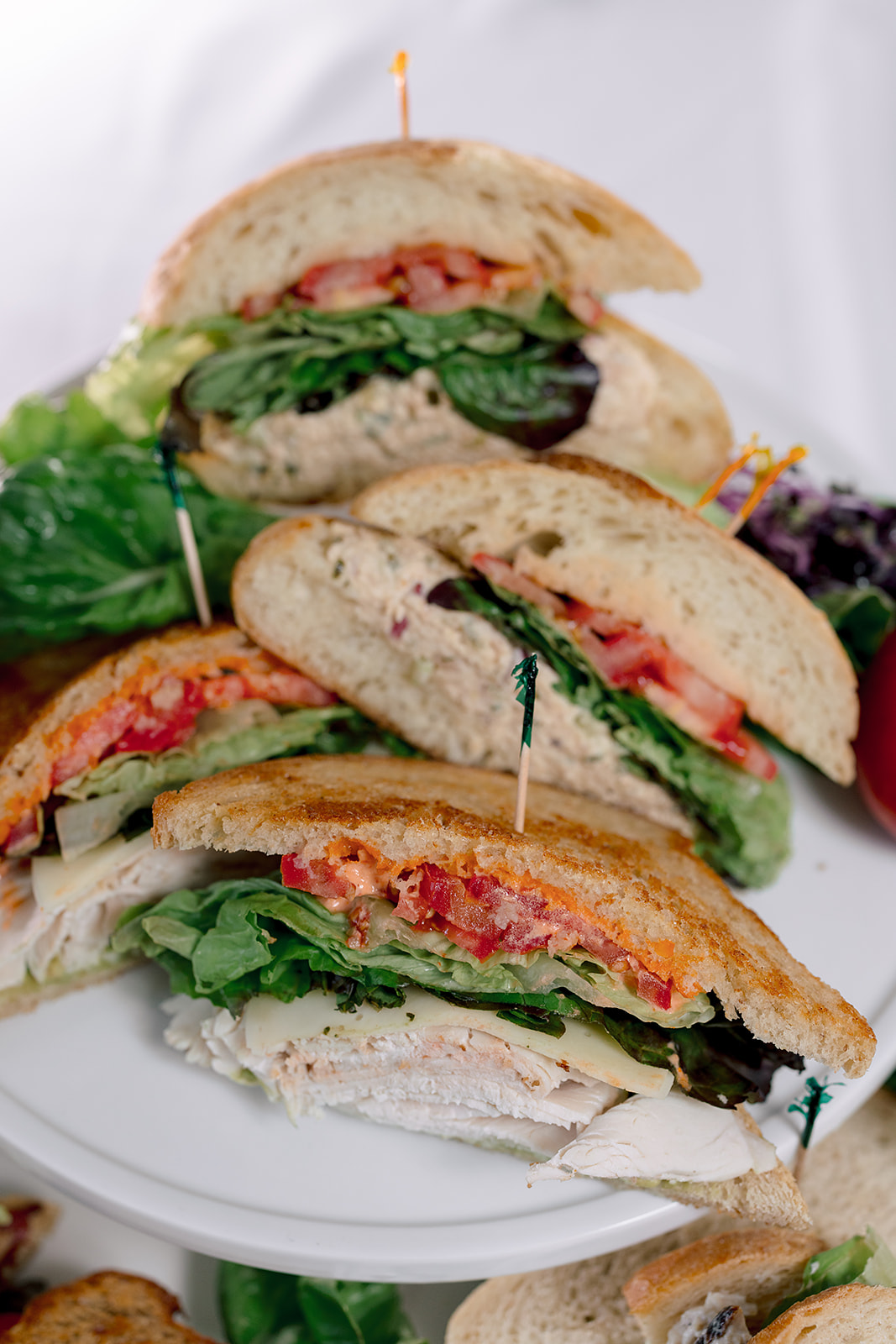 Stacked sandwiches with bread, lettuce, tomato, and meat on a white plate, with a blurred background.