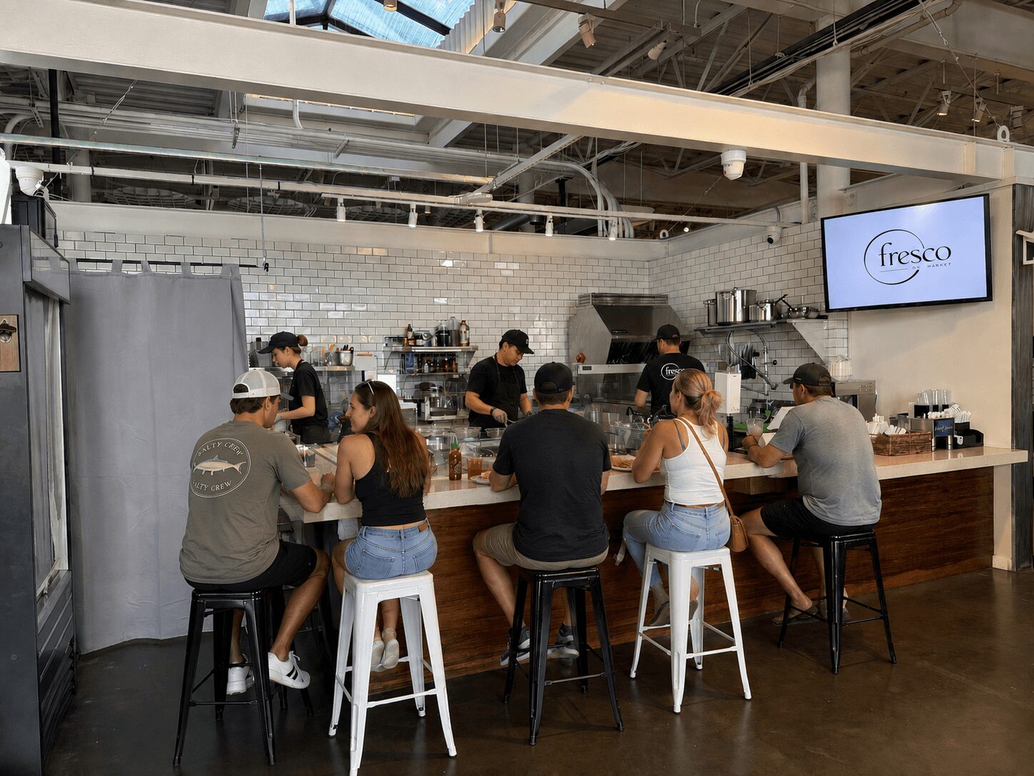 People sitting at a bar counter in a cafe, with a kitchen area and a screen displaying 'fresco' in the background.