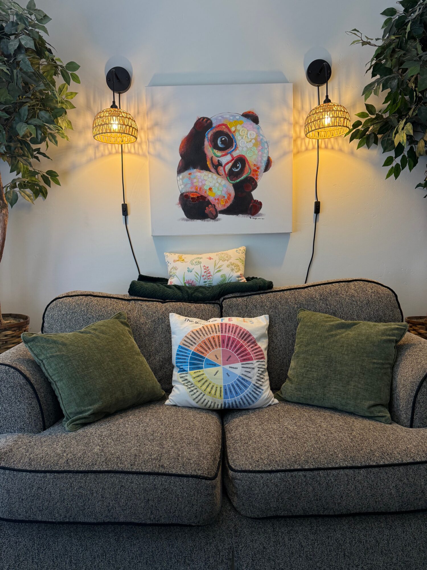 Living room with a gray sofa, colorful pillow, panda artwork, and two hanging lamps, surrounded by green plants.