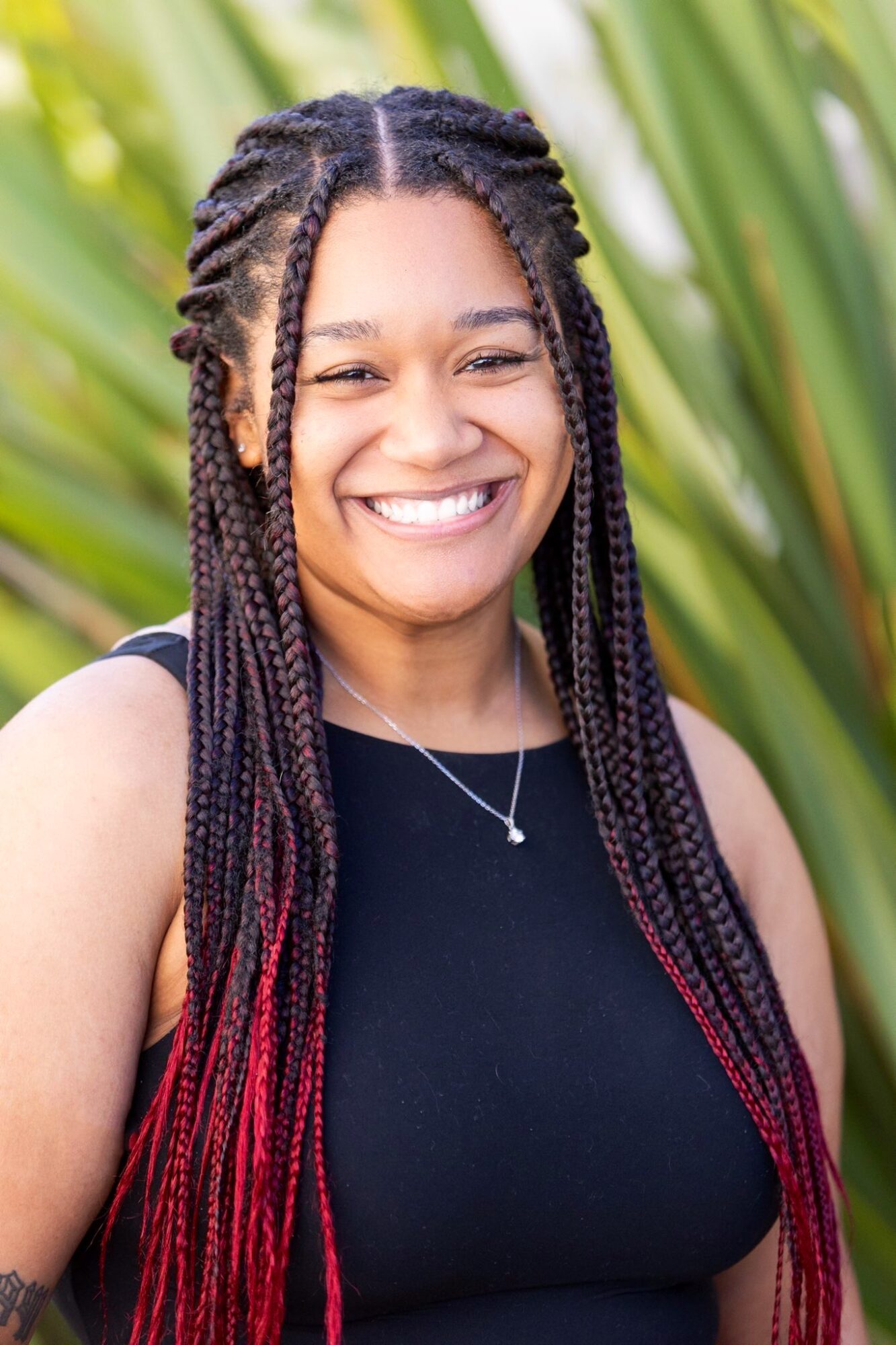 Smiling woman with braided hair, wearing a black top and necklace, outdoors with green plants in background.