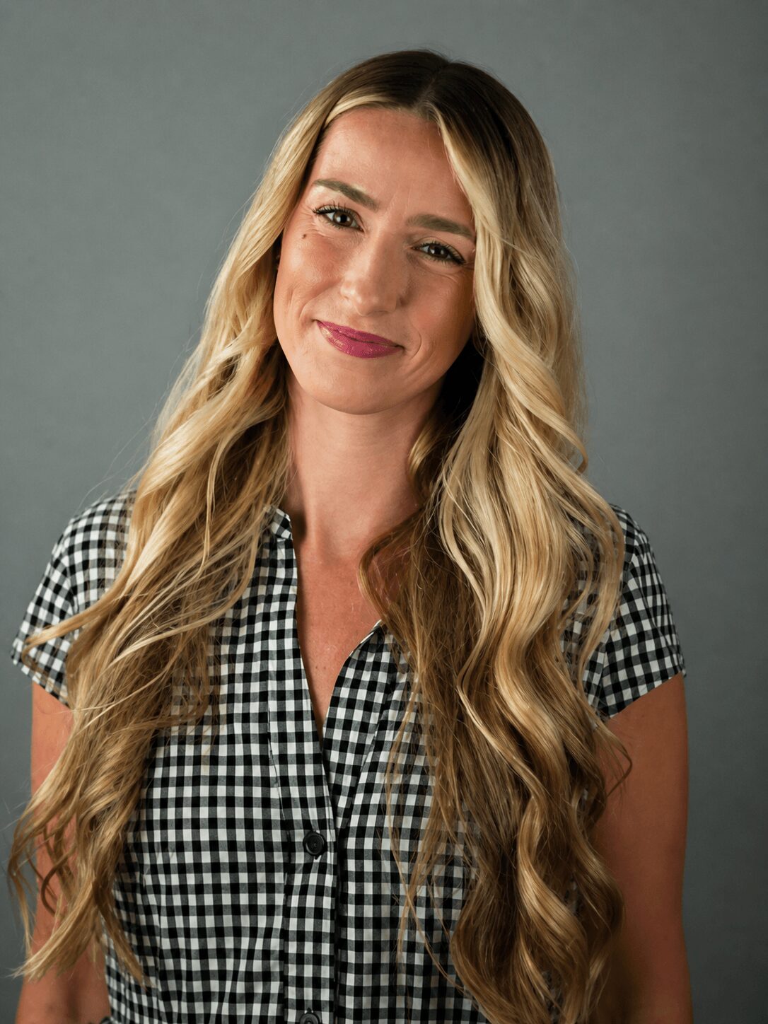 Woman with long wavy blonde hair wearing a black and white checkered shirt, smiling softly, against a plain gray background.