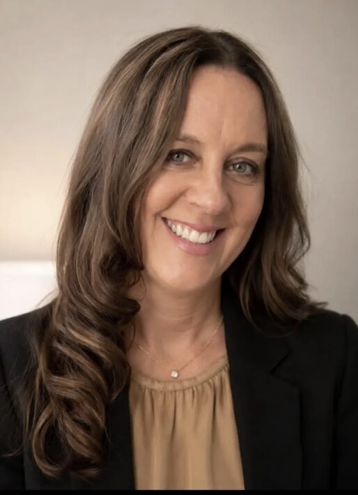 Woman with long wavy hair smiling, wearing a black blazer and beige top, neutral background.