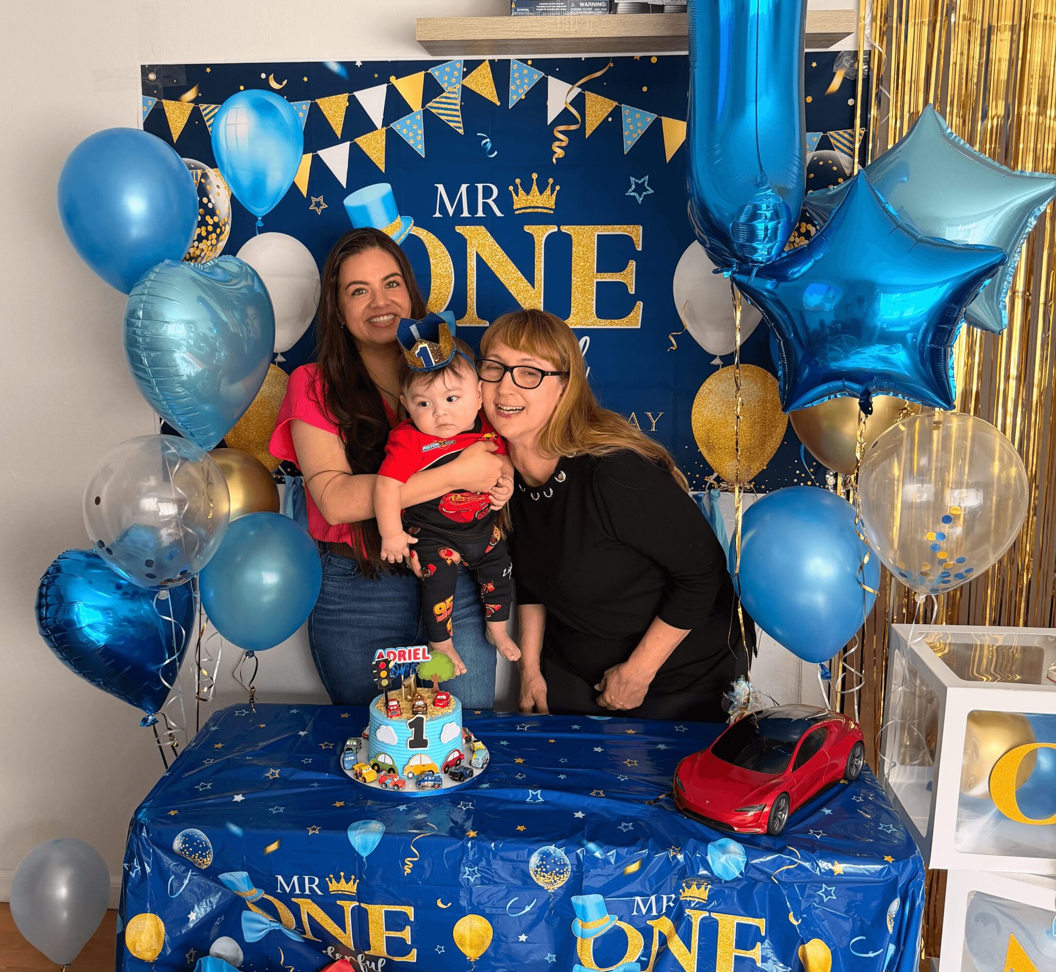 Two women and a child celebrating a first birthday with balloons, a cake, and a red toy car, in front of a decorated backdrop.