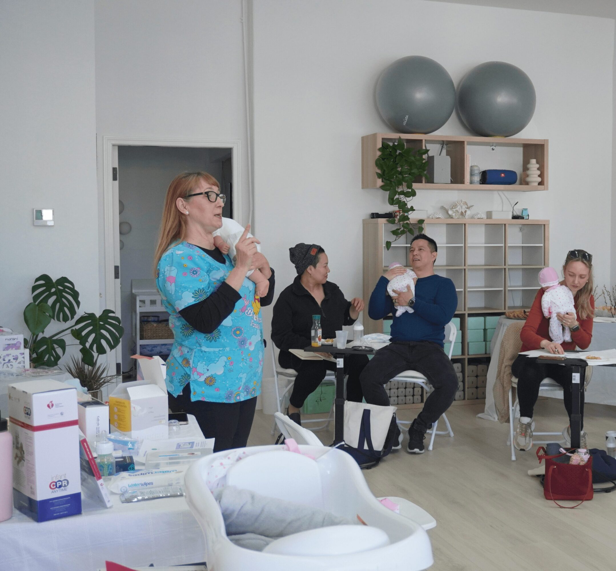 Group of women and children in a room, some holding babies, with shelves and exercise balls in the background.
