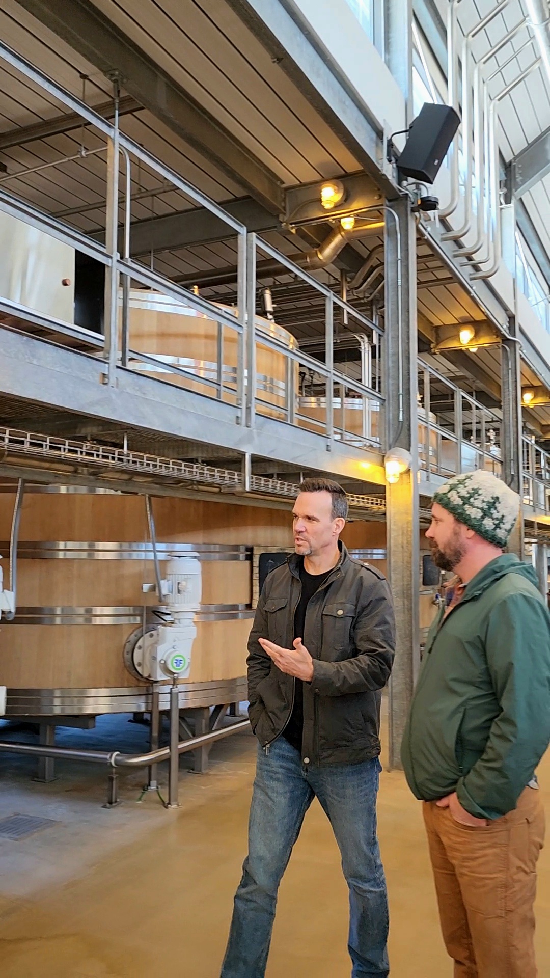 Two men stand and talk inside a brewery with large tanks and metal railings, one gesturing with his hand.