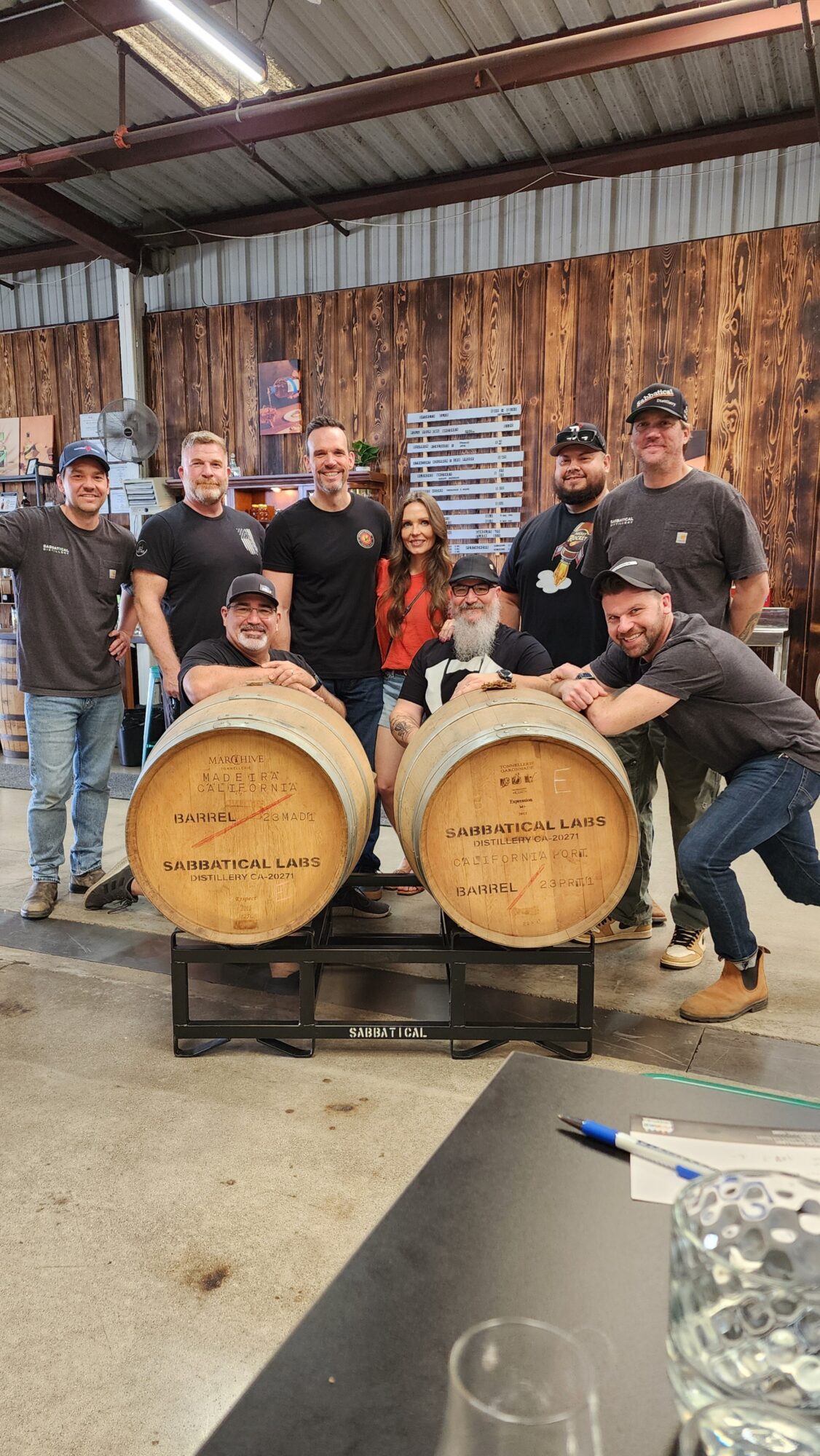 Group of seven people standing behind two wooden barrels on a stand, in a rustic indoor setting with wood-paneled walls.