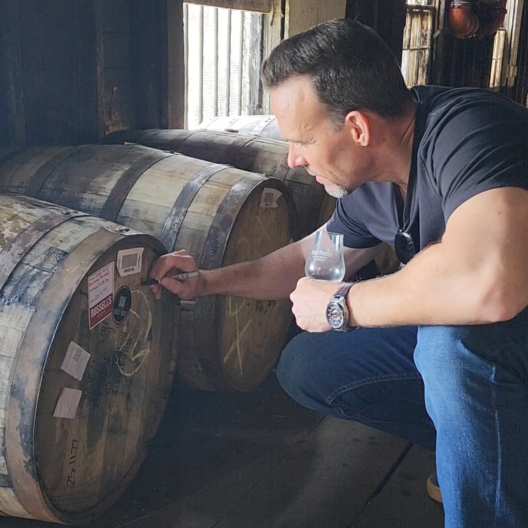 Man inspecting barrels in a rustic setting, holding a glass and pointing at a label on a barrel.