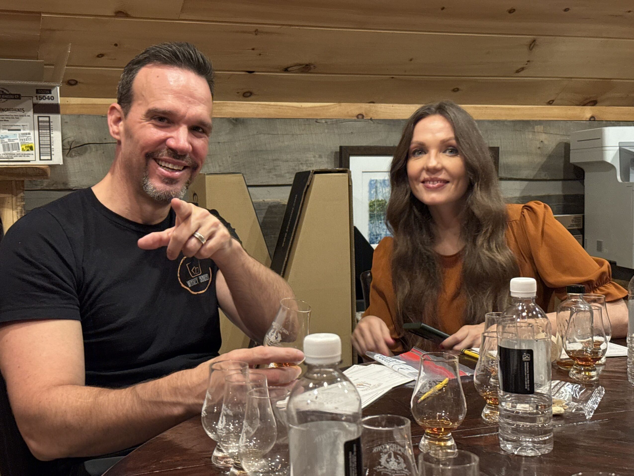 Two people sitting at a table with drinks, smiling, in a room with wooden ceiling and concrete wall.