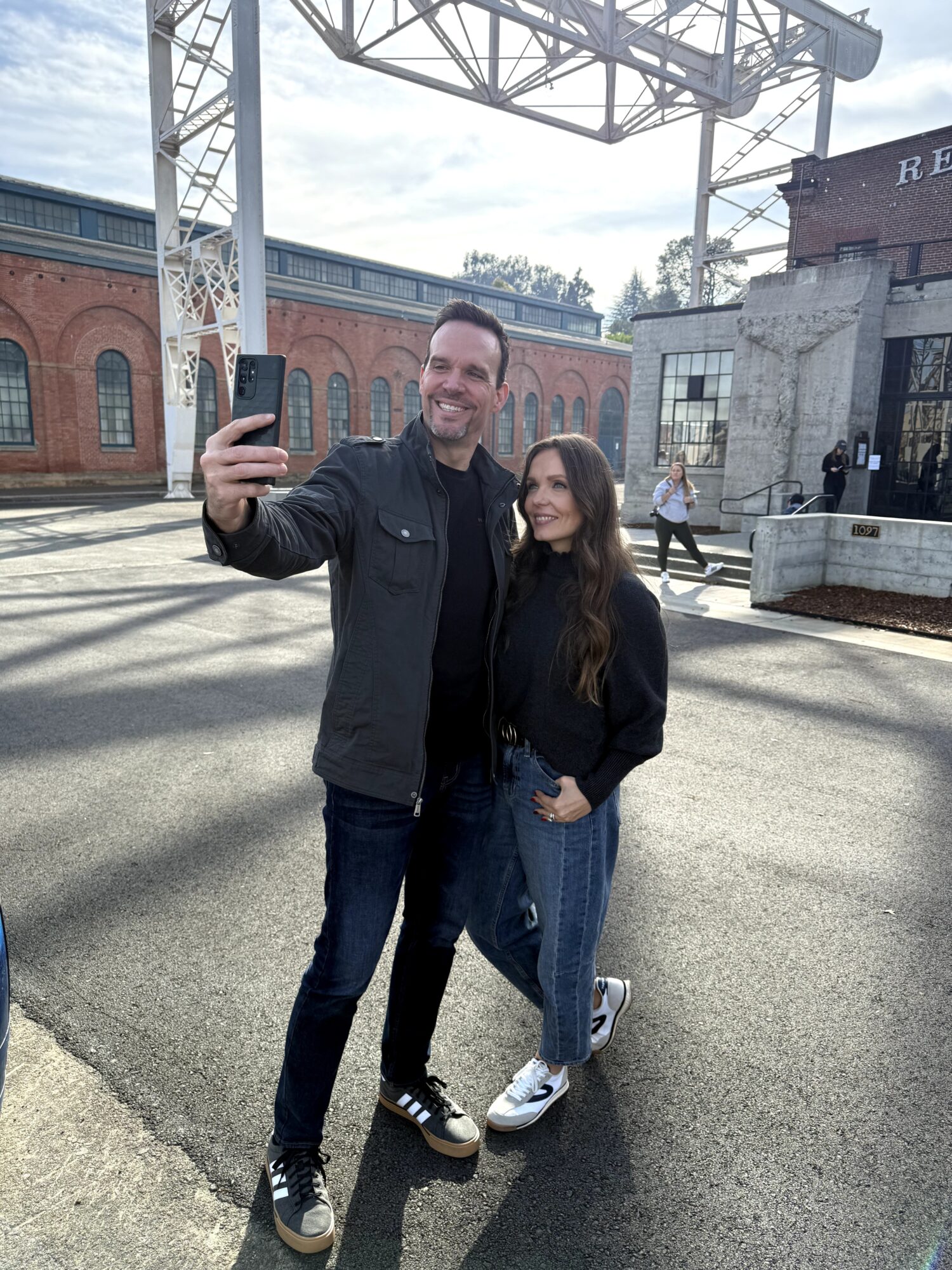 Man and woman taking a selfie outdoors with industrial buildings in background.