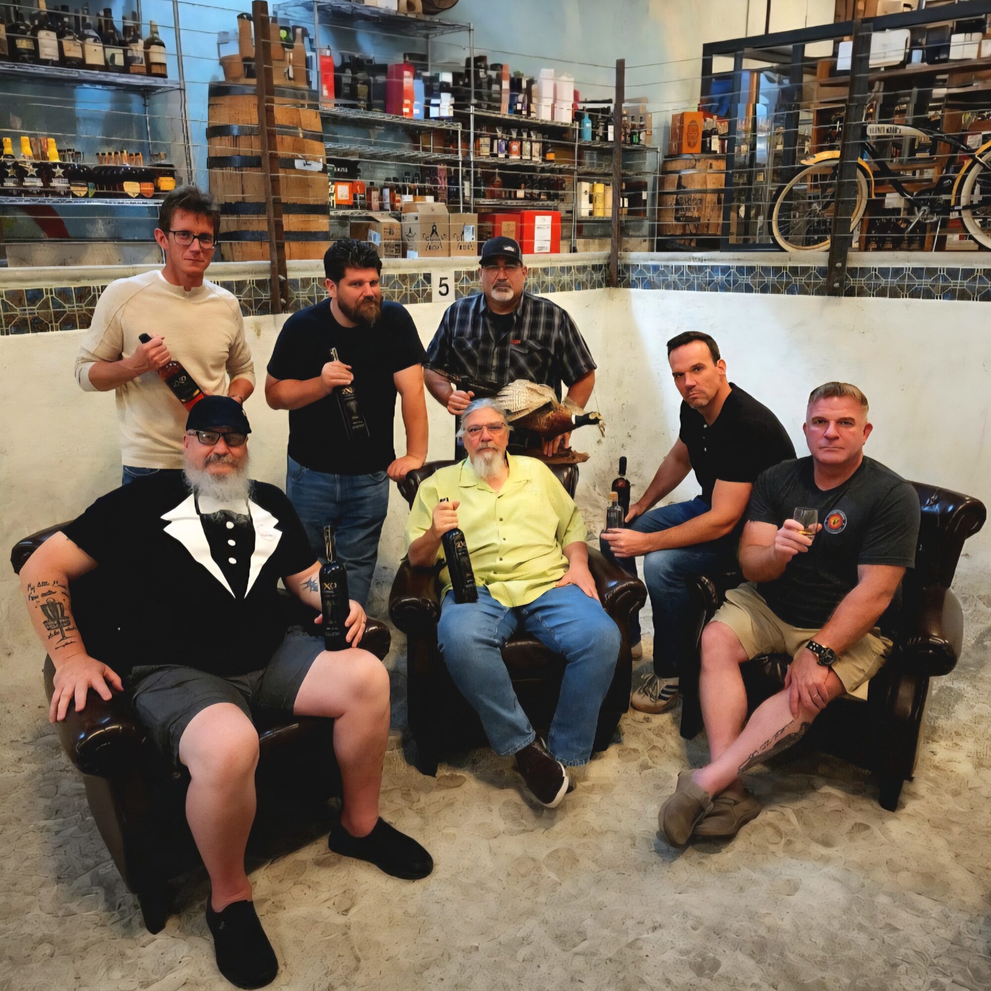 Group of seven men sitting and standing in a warehouse, some holding bottles, with shelves and bicycles in background.