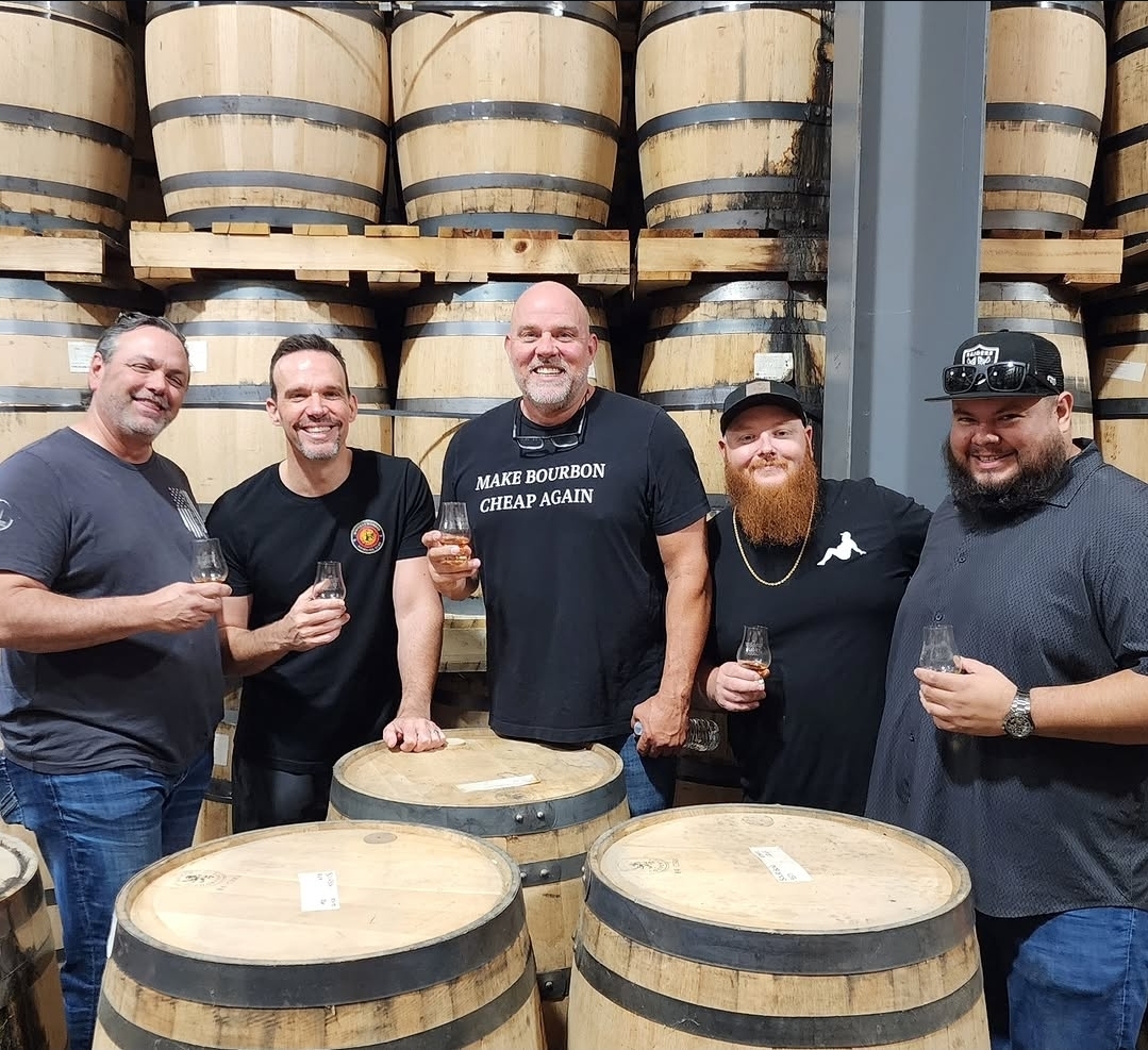 Five men standing in a warehouse with stacked barrels, holding glasses, smiling, and posing for a photo.