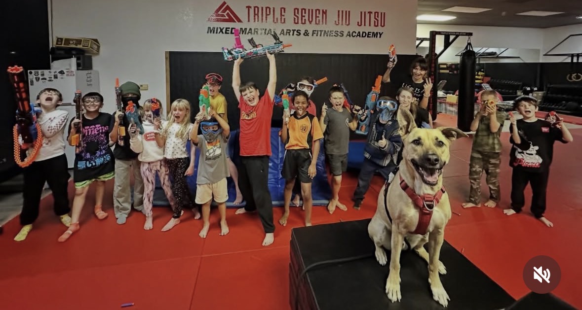 Group of children and adults in a martial arts gym, with a dog in the foreground, on a red mat.