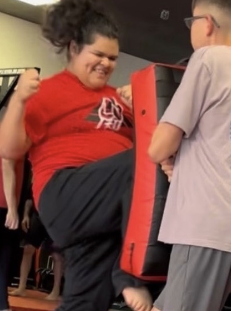 Child in red shirt appears to be kicking or raising leg, with another child in gray shirt nearby, in an indoor setting.