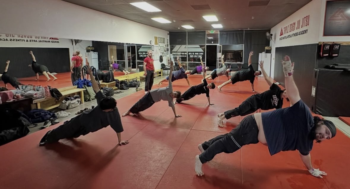 Group of people doing side plank exercises on red mats in a gym, with equipment and mirrors in the background.