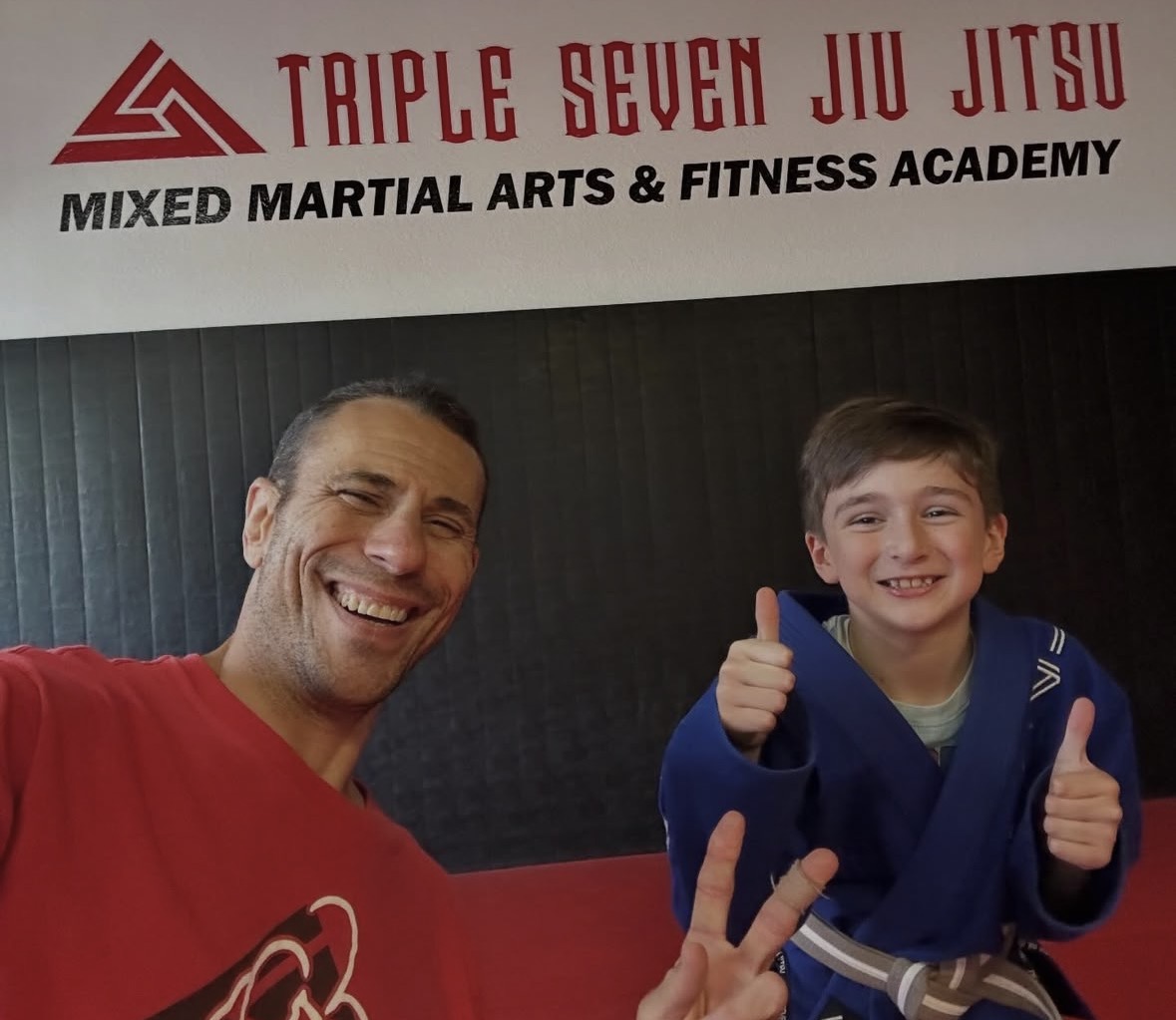 Two people smiling and giving thumbs up in a martial arts gym with a sign reading 'Triple Seven Jiu Jitsu'.