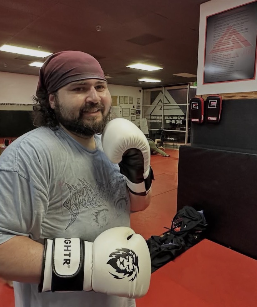 Man with a beard and headscarf in boxing gloves at gym, smiling, holding gloves near face, background with gym equipment.