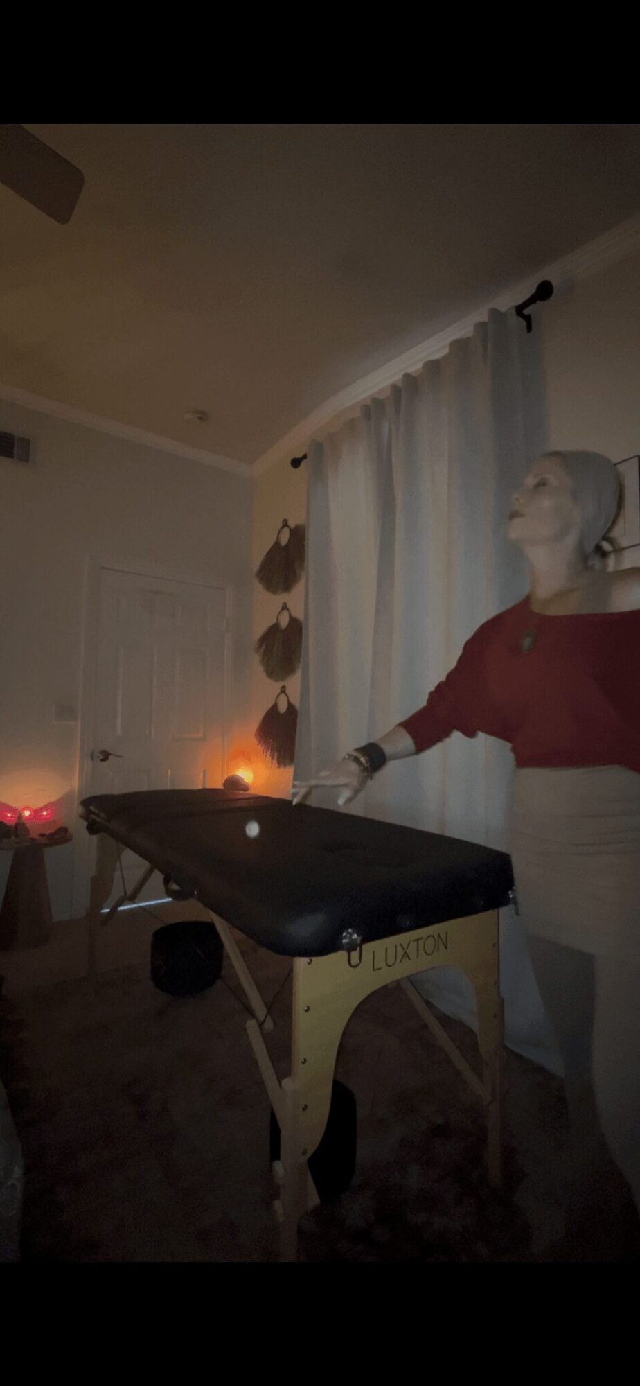 Person wearing a mask and red sweater stands near a table with a laptop, in a dimly lit room with curtains.
