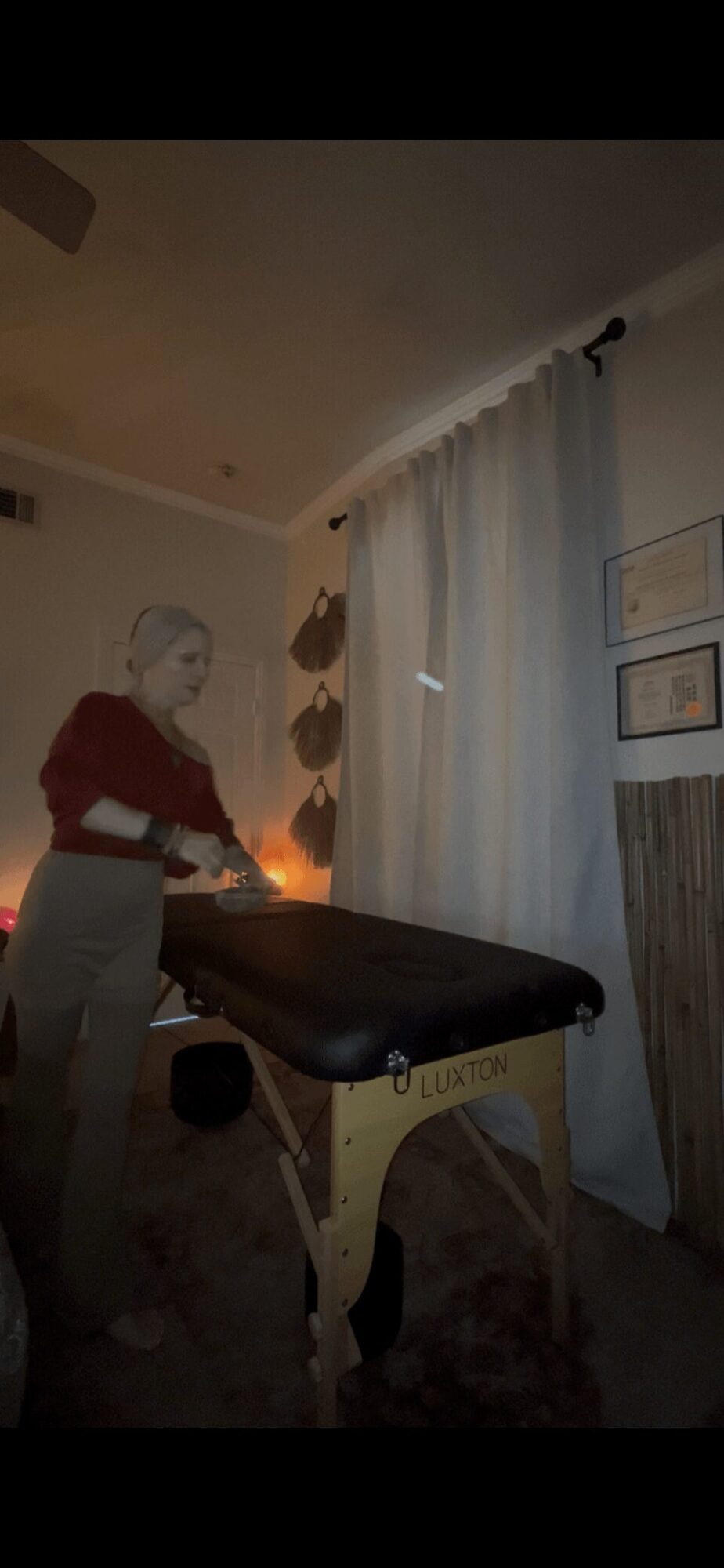 Person with short hair in red top and gray pants stands beside a massage table in a dimly lit room with curtains and framed pictures.