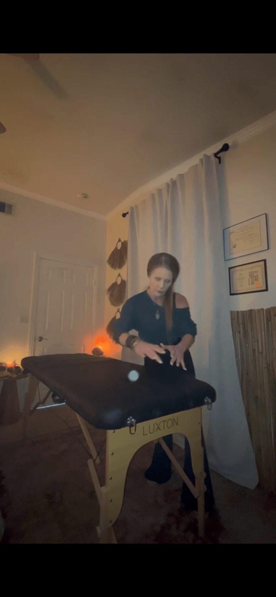 Person standing at a massage table in a room with curtains and framed pictures on the wall.