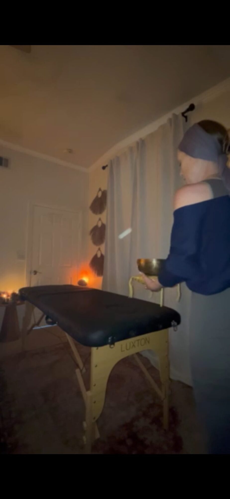 Person with blindfold holding a bowl, standing next to a massage table in a dimly lit room with curtains.