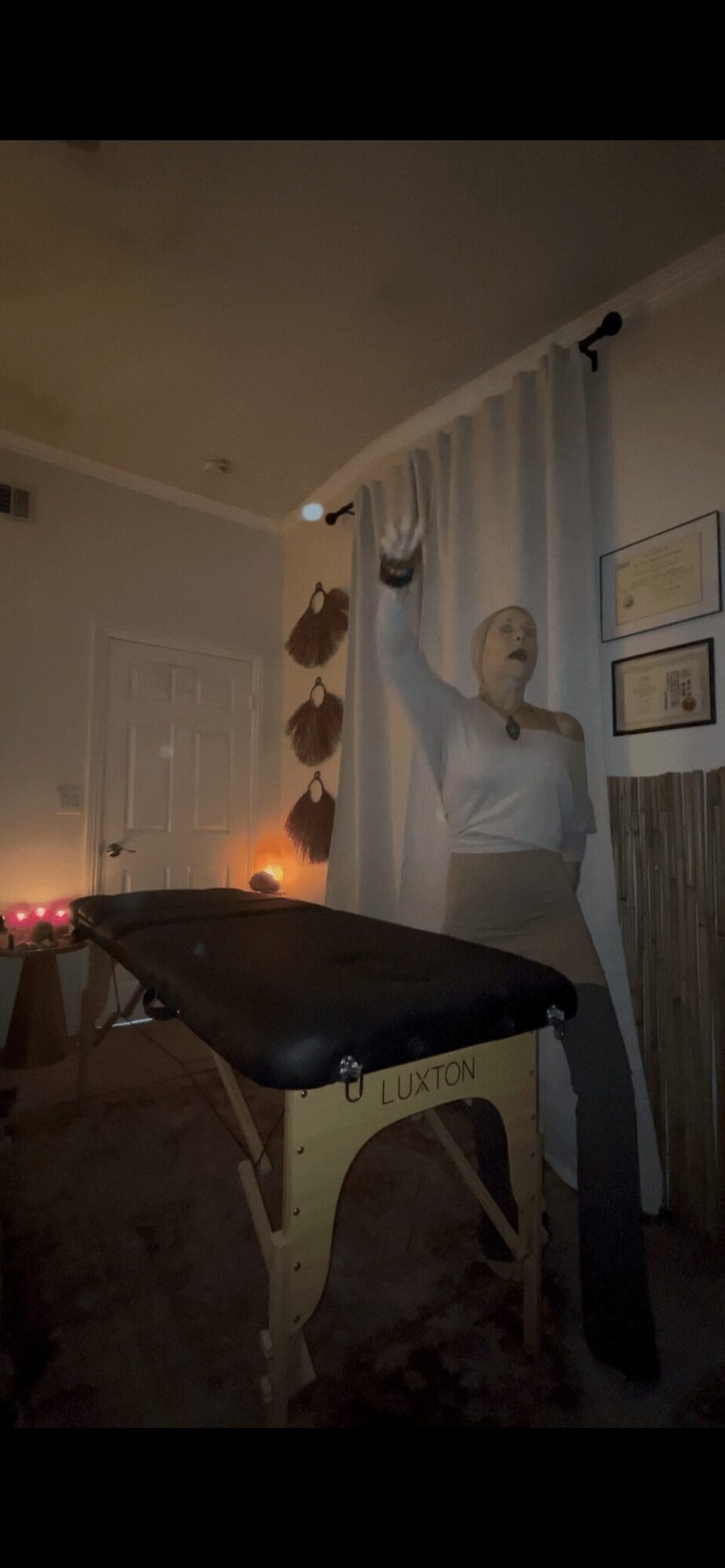 Person standing next to a curtain, reaching up with one hand, in a dimly lit room with framed pictures on the wall.
