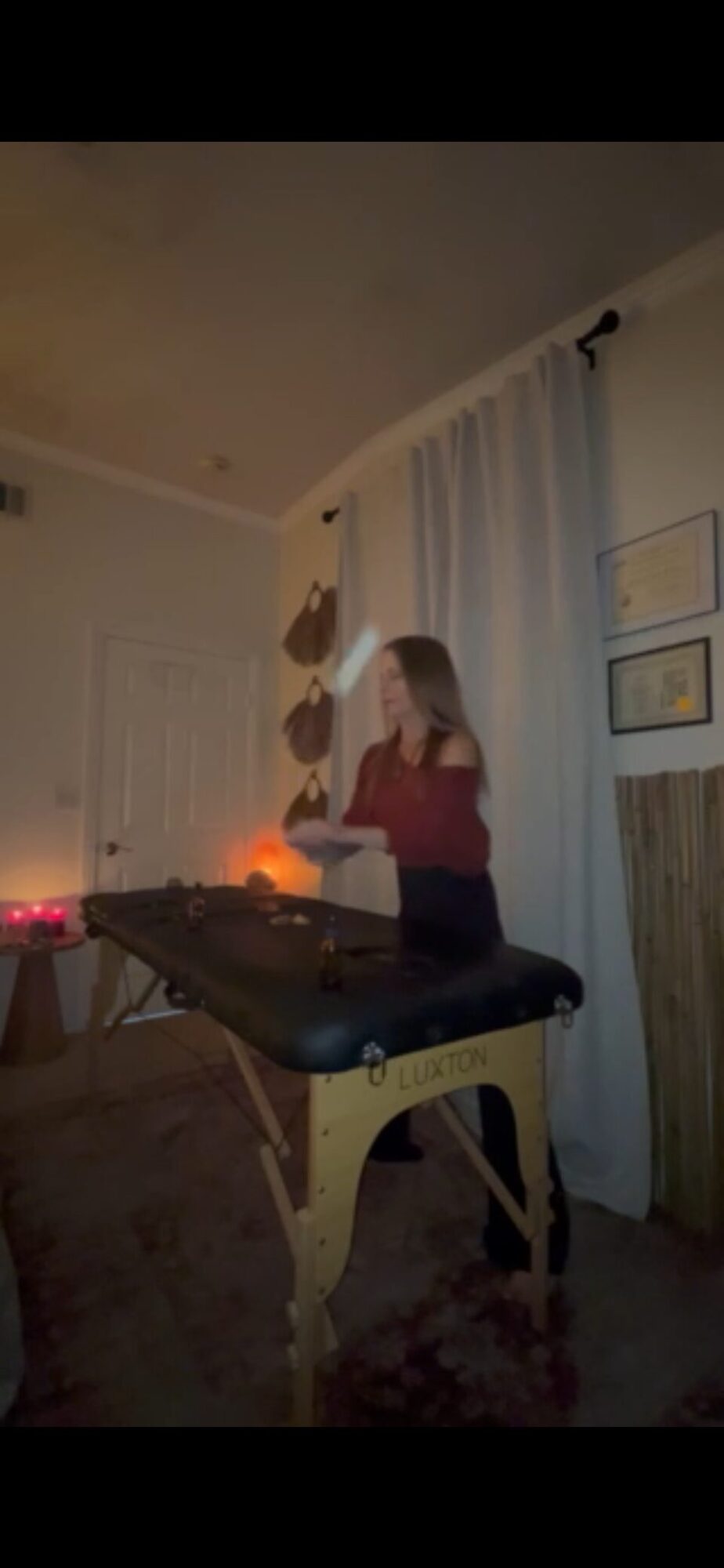 Person standing at a table with a candle, in a room with curtains and framed pictures on the wall.
