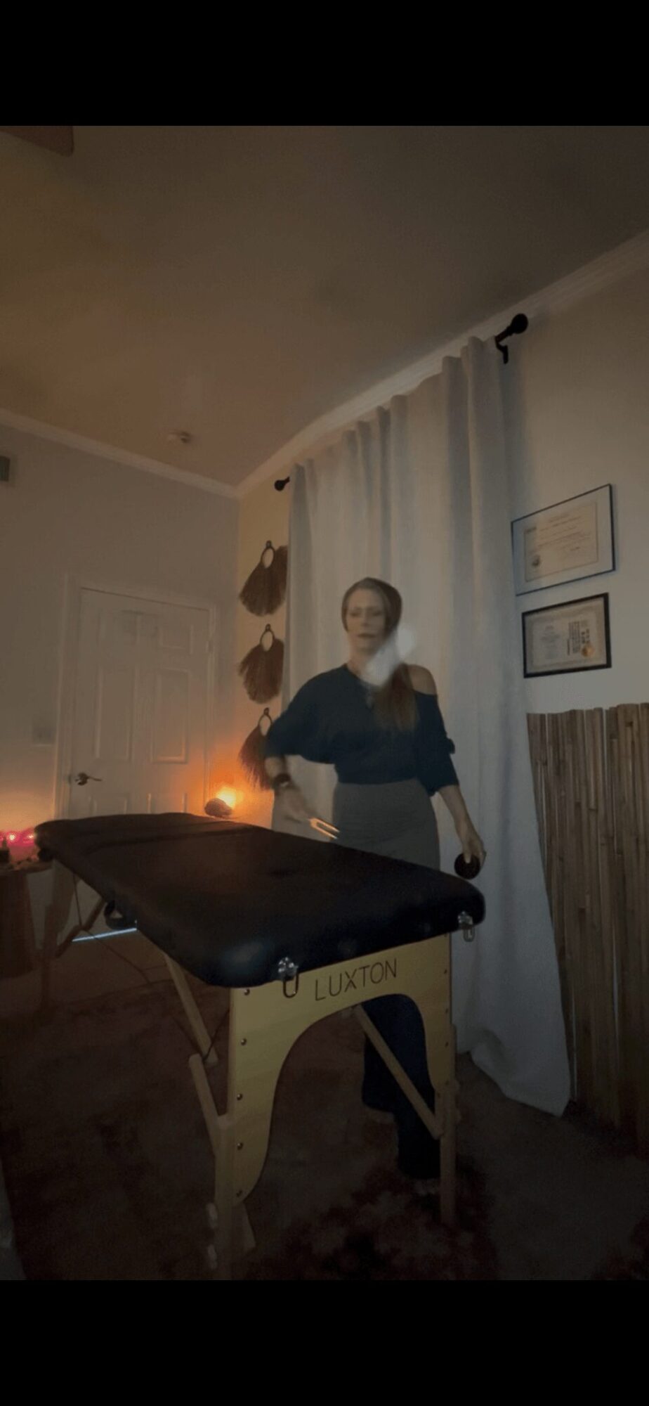 Person standing next to a massage table in a room with curtains and framed pictures on the wall.
