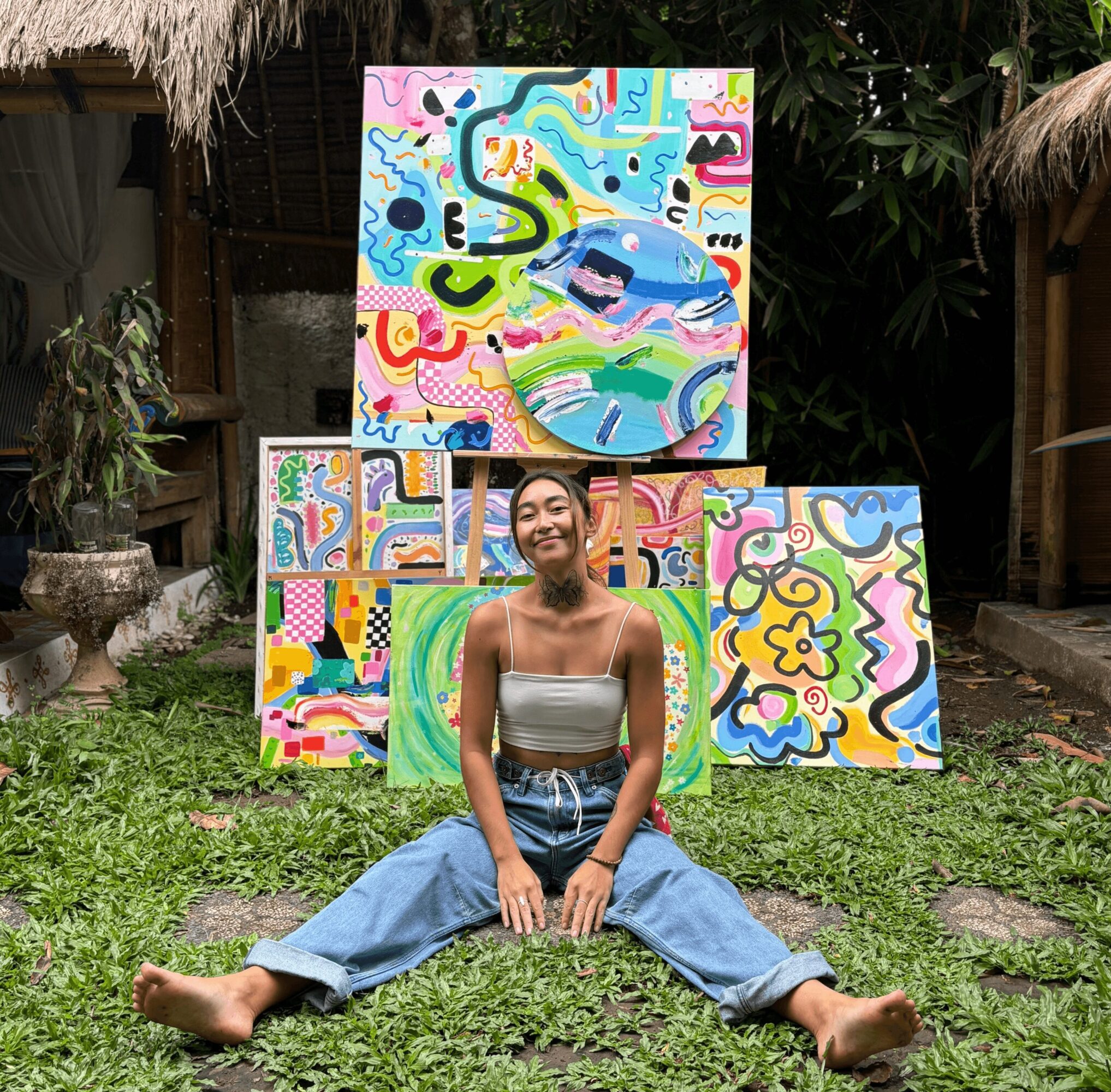 Person sitting on grass with legs spread, smiling, in front of colorful abstract paintings outdoors.