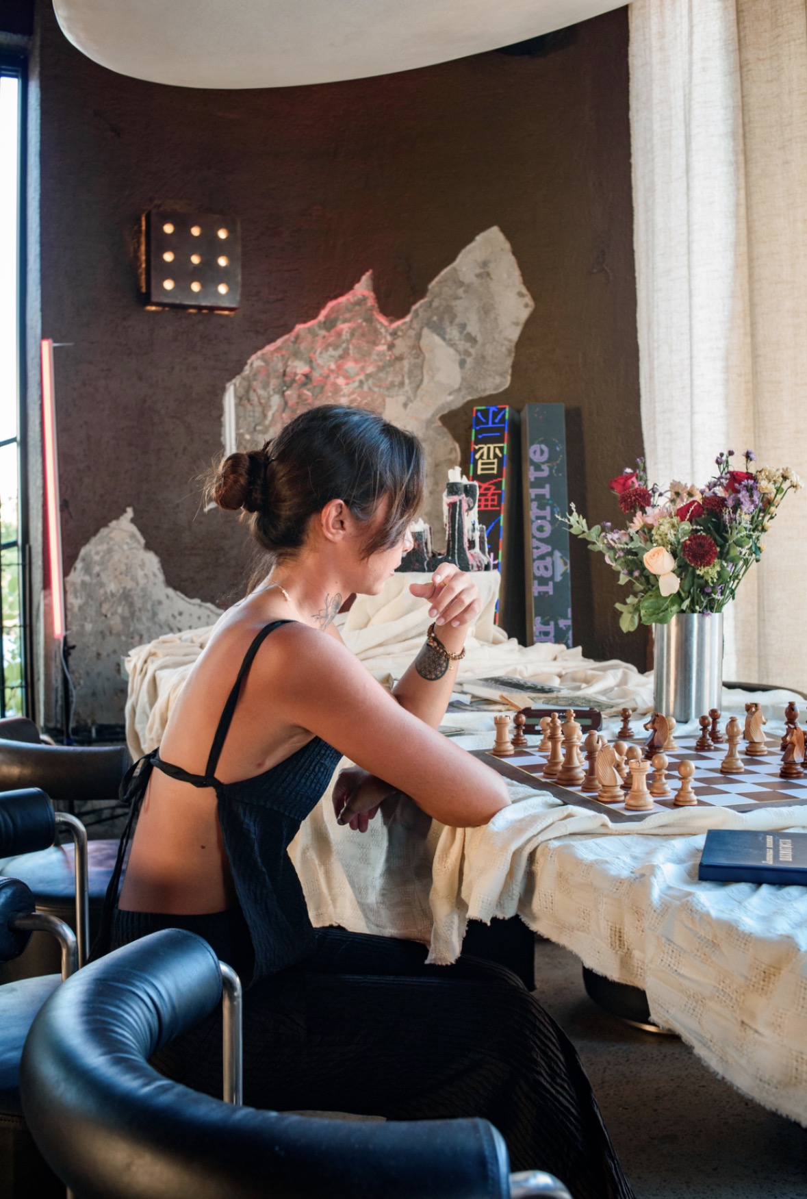 Woman with dark hair in a bun, wearing a black dress, playing chess at a table with a flower arrangement.