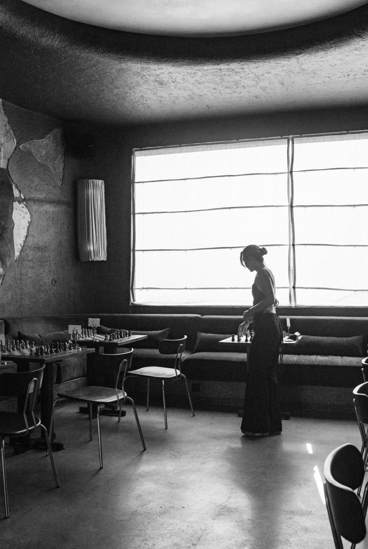 A woman stands near a large window in a restaurant, with tables and chairs around her, in black and white.