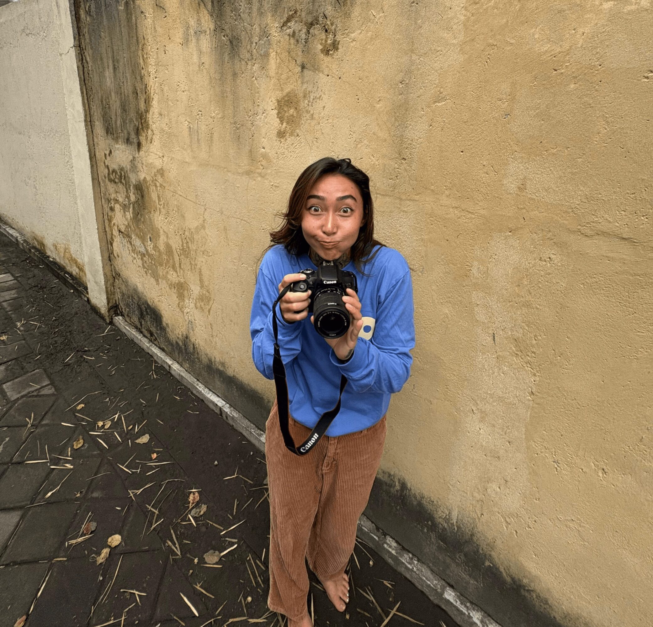 Person with shoulder-length hair wearing a blue shirt and brown pants, holding a camera, standing against a yellow wall.