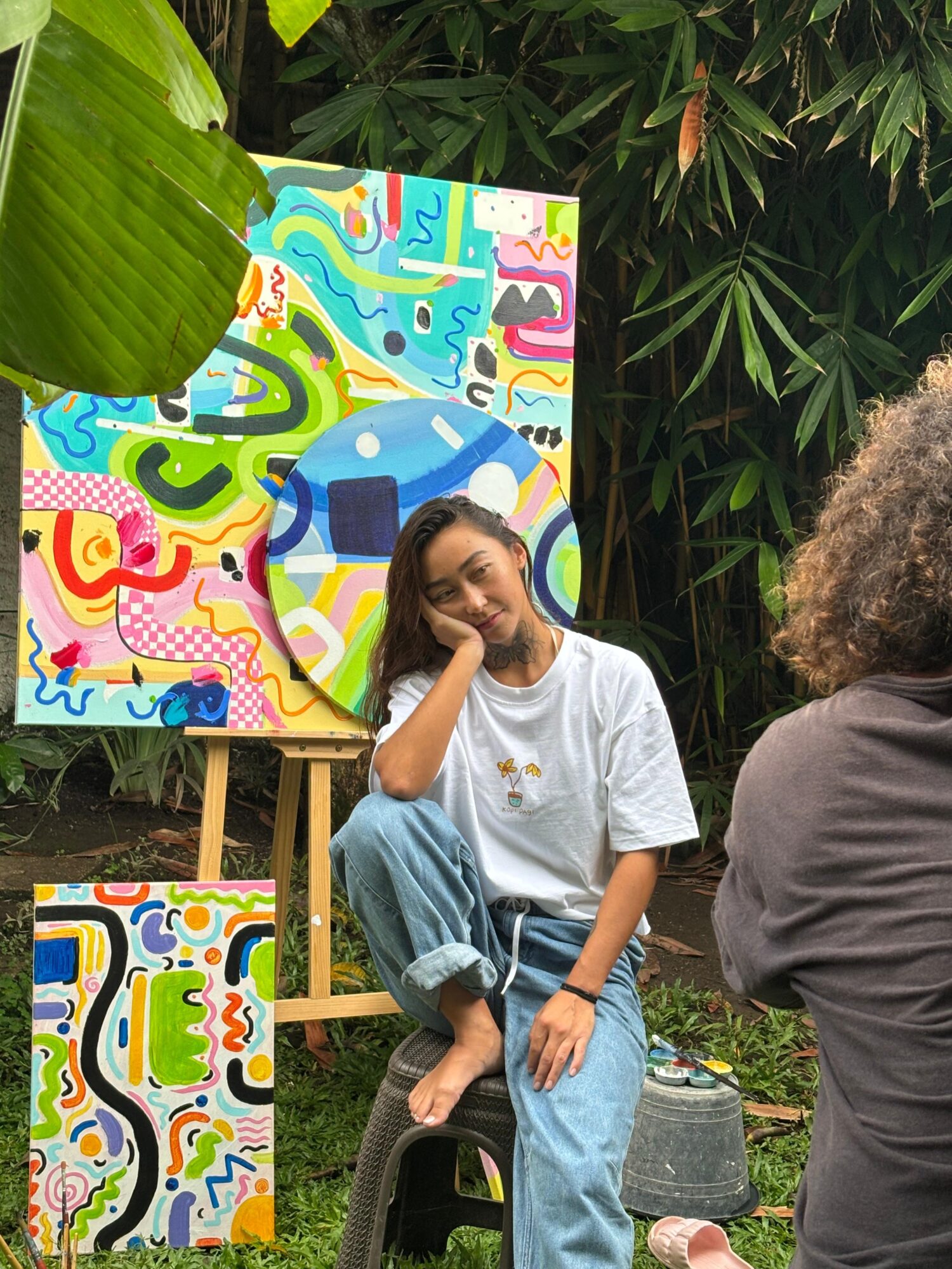 Young woman sitting on a stool in front of colorful abstract paintings outdoors, smiling and resting her head on her hand.