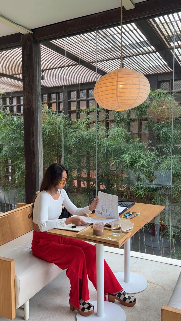 Woman sitting at a table with a laptop, in a bright room with large windows and hanging paper lantern, greenery outside.