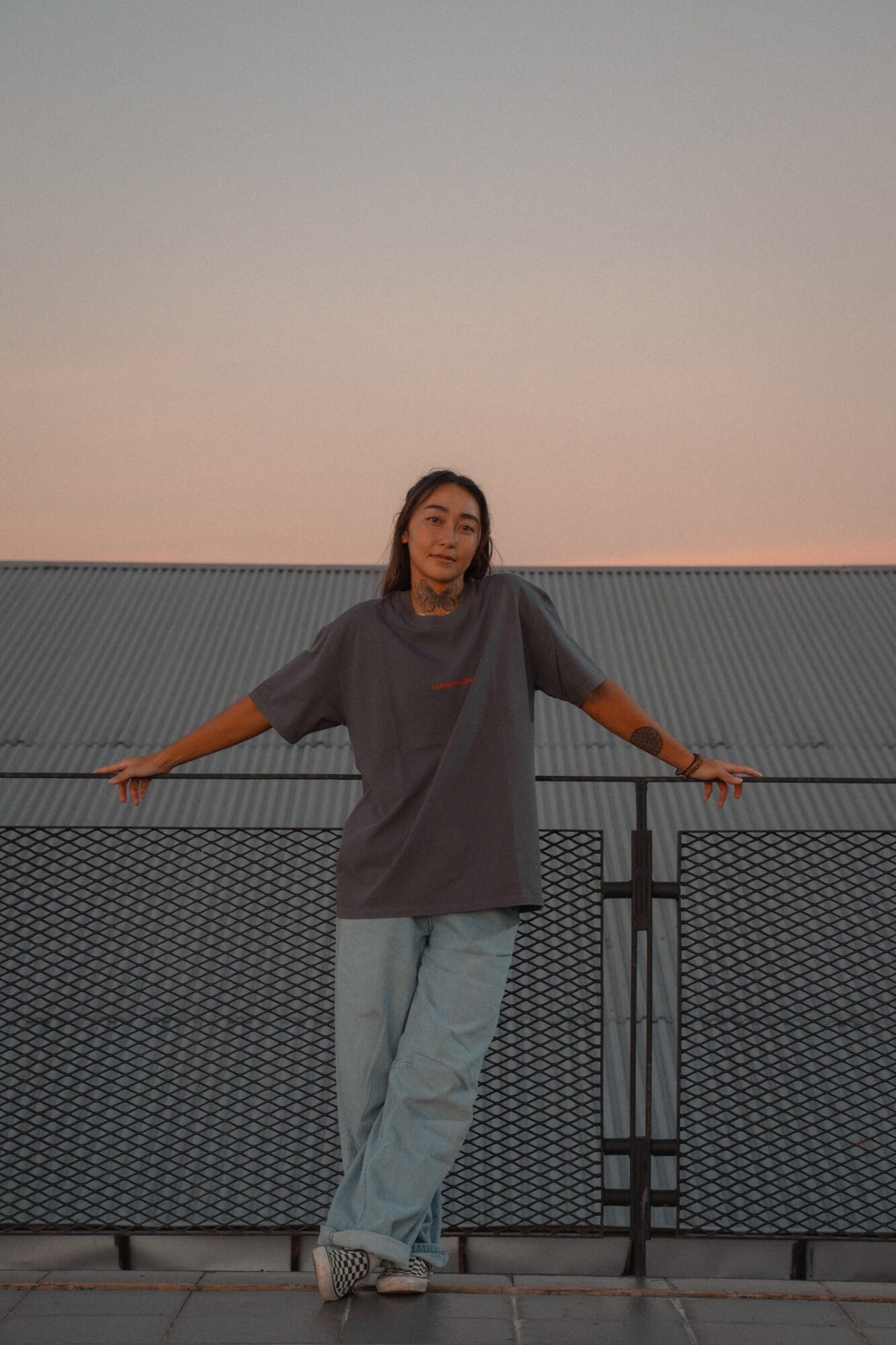 Person standing on a rooftop with arms outstretched, wearing a loose gray shirt and light jeans, against a sunset sky.